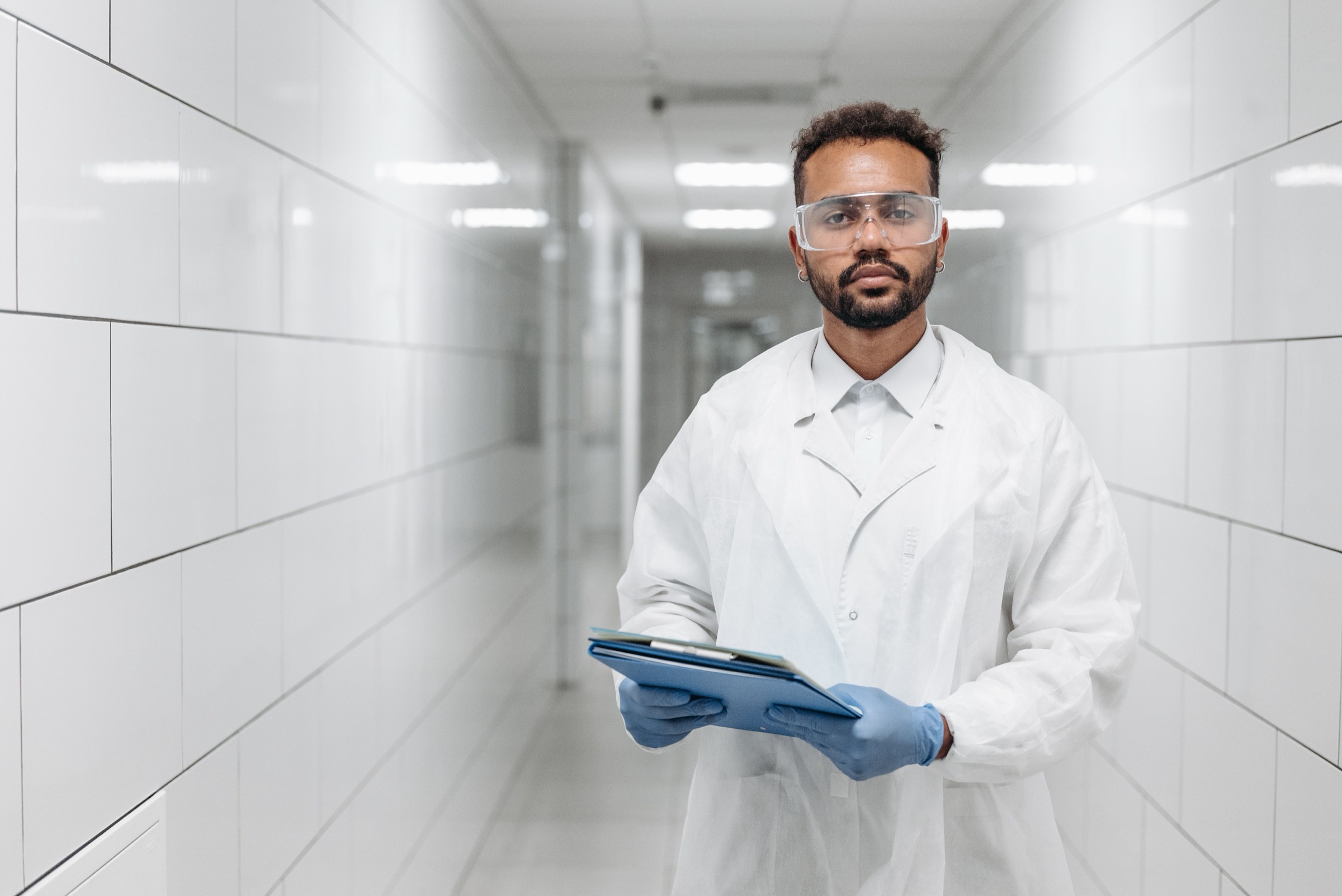 An operations leader in a white coat and safety glasses standing in a plant corridor, holding a tablet and looking at the camera