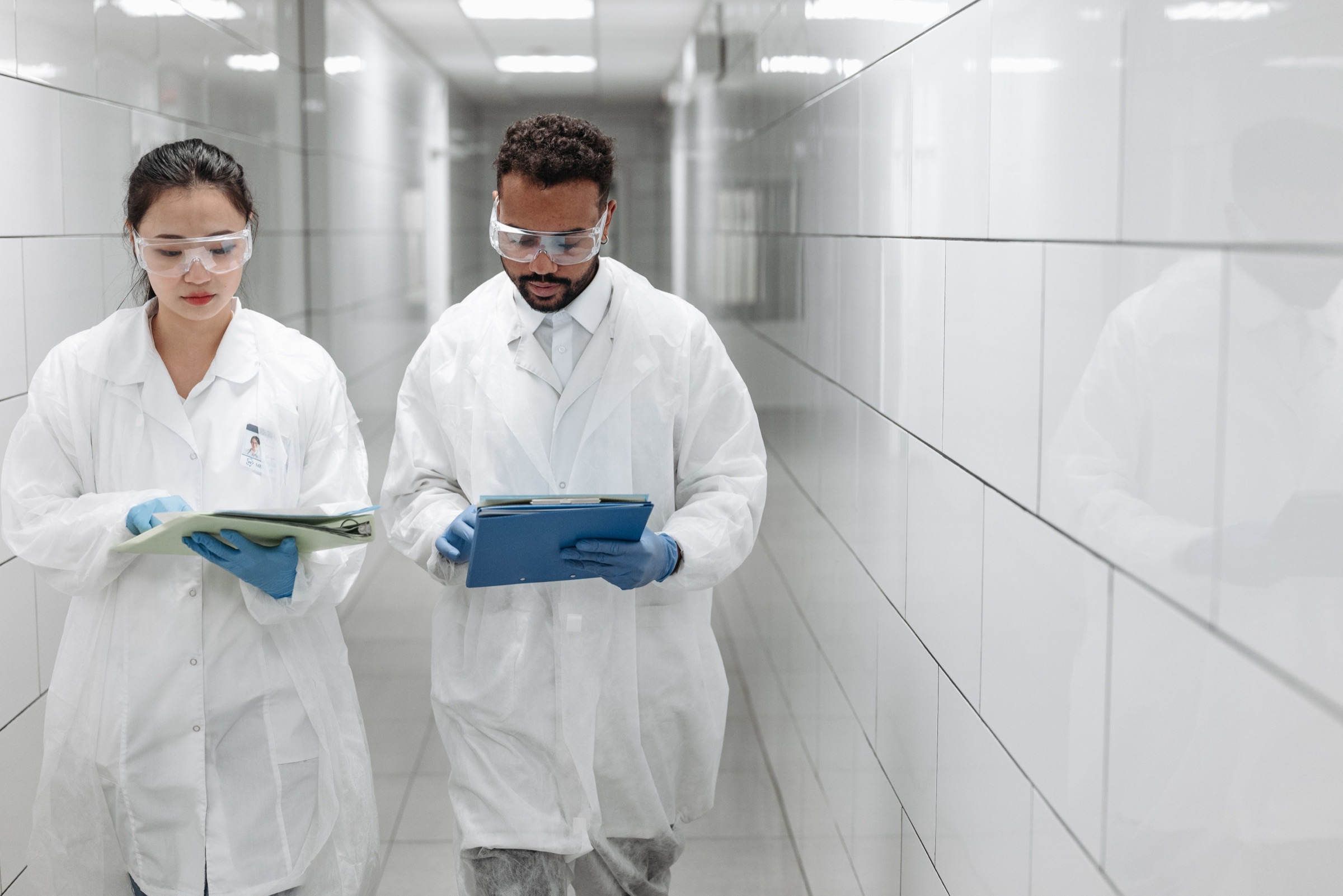 Two operations leaders in white coats and safety glasses walking a plant corridor, reviewing a tablet and a clipboard